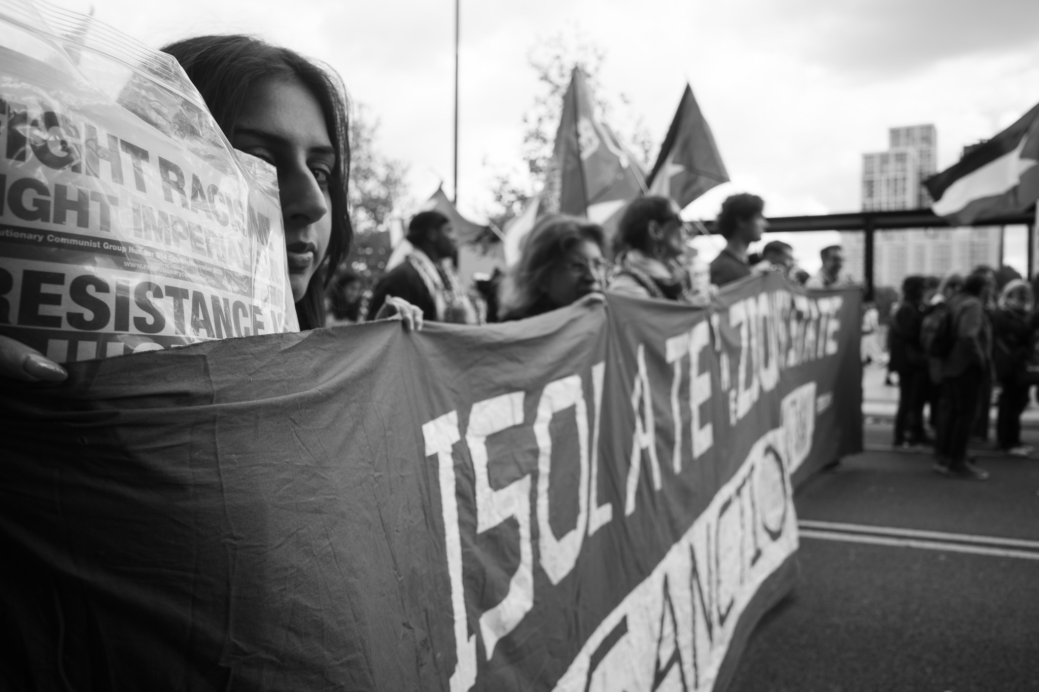 A young woman half hidden behind a banner and a plastic-wrapped newspaper, marchers and flags behind her