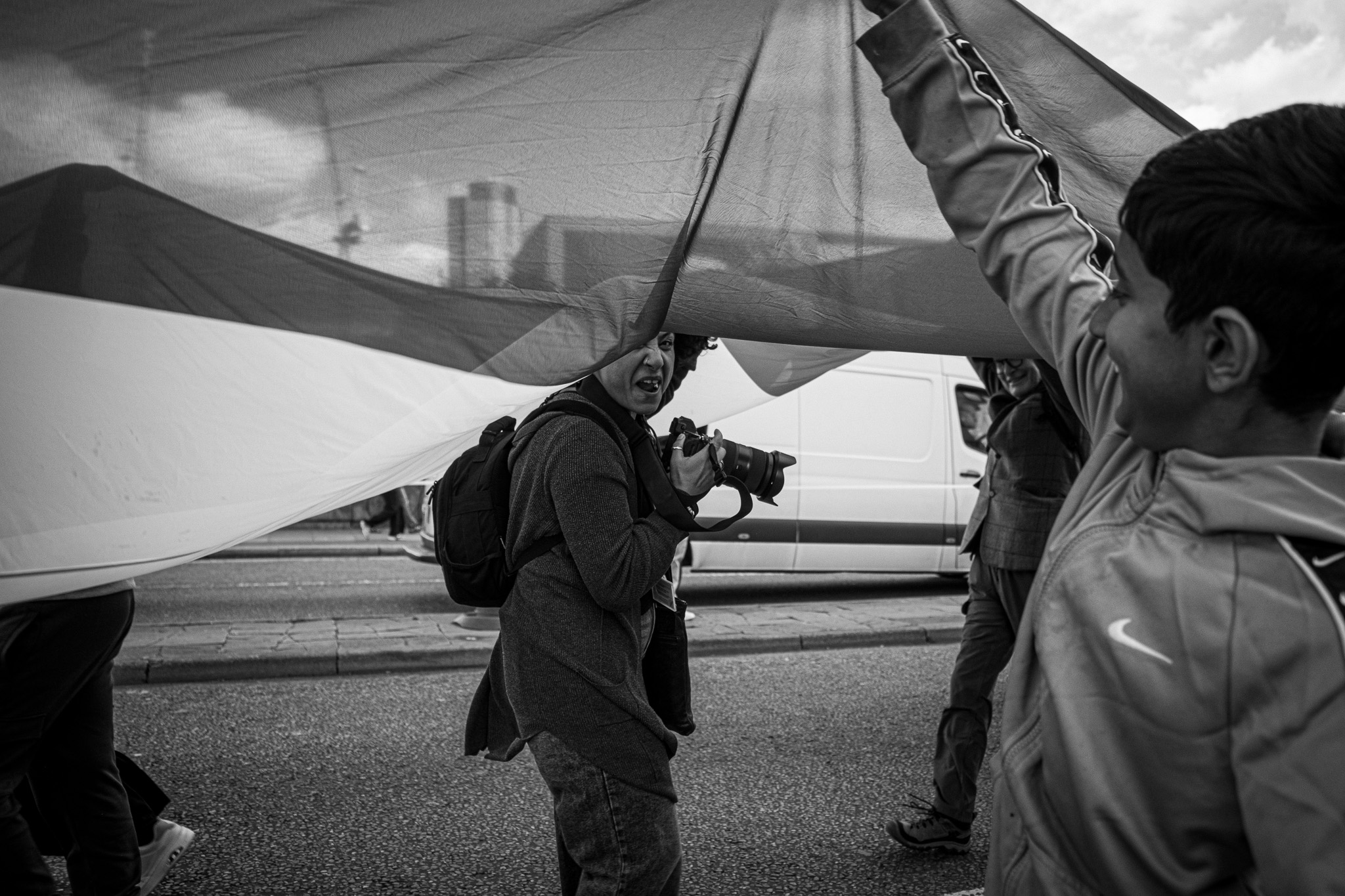 A photographer with a camera glancing back at the viewer from under a large translucent flag, a smiling boy at the right edge