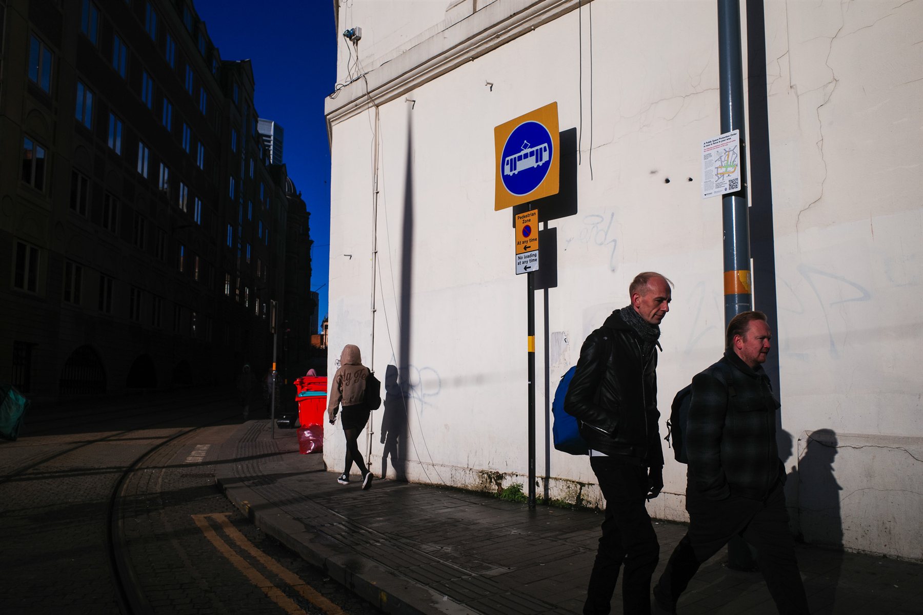 Two men walking along a sunlit white wall with long shadows
