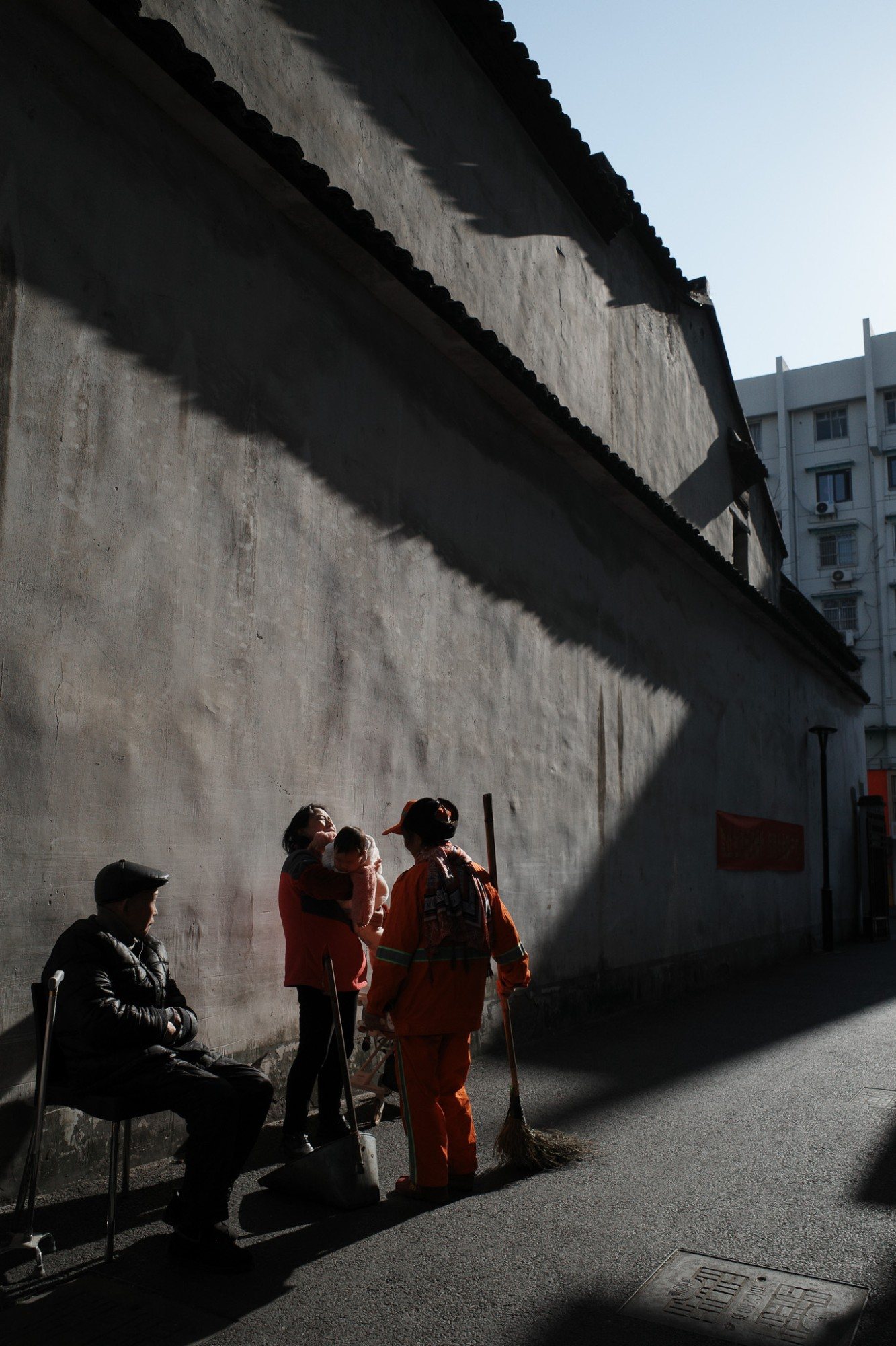 Street sweepers and a woman holding a baby in a diagonal band of light against a long wall