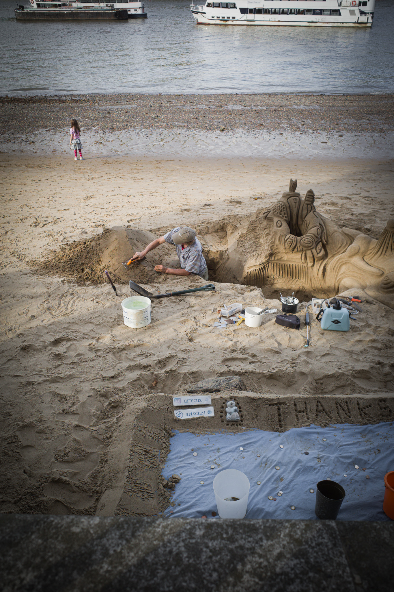 A man digging into a beach to carve a sand sculpture while a girl stands at the water's edge